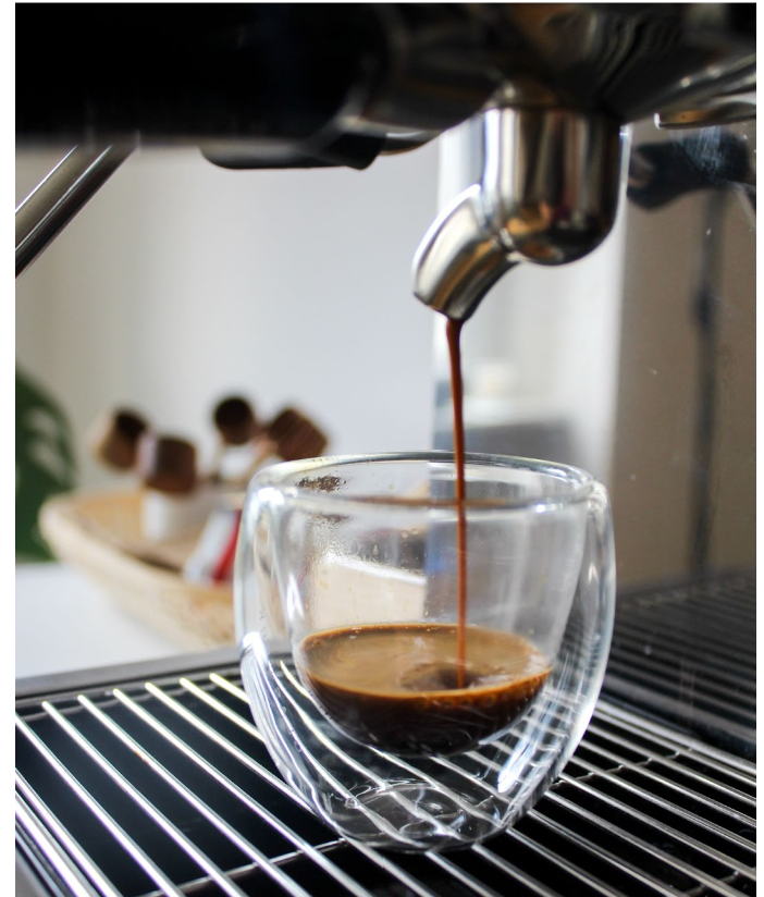 Espresso being poured into a clear glass cup from an espresso machine, with a basket of plants in the background.