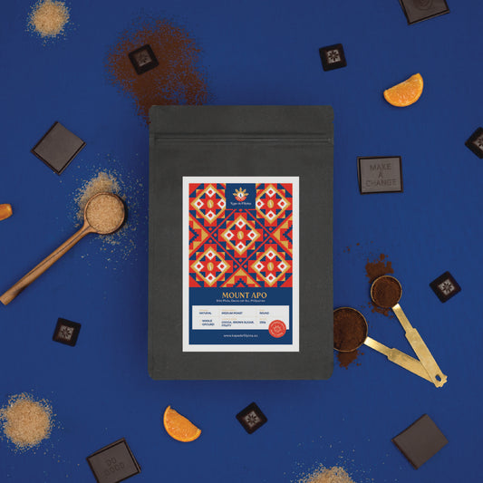 An Arabica coffee bag on a blue background surrounded by coffee beans, ground coffee, and chocolate pieces.